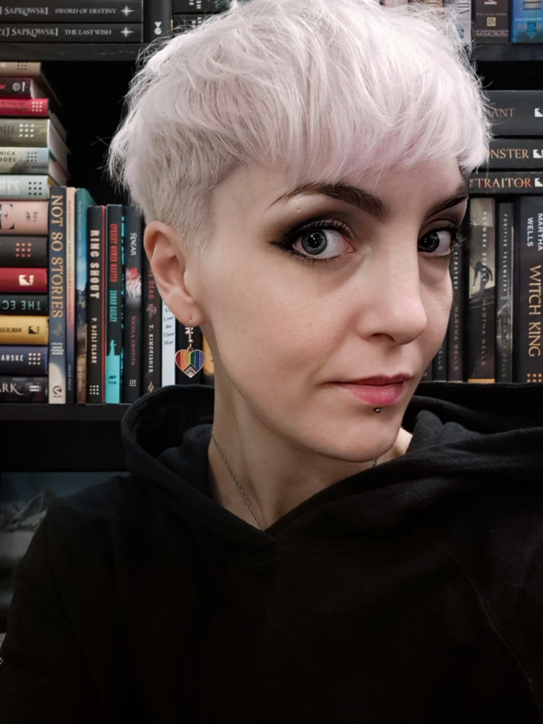 A photo of Liv, a fair-skinned enby with platinum-colored hair cut into a gender neutral pixie. Liv is wearing Pride flag earring and a black hoodie, and is pictured in front of a bookshelf full of titles from Tor and Tordotcom. Photo used for The Best Book Nook.